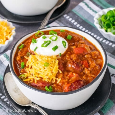 A white bowl filled with Dr. Pepper Chili and topped with sour cream, shredded cheese, and green onions