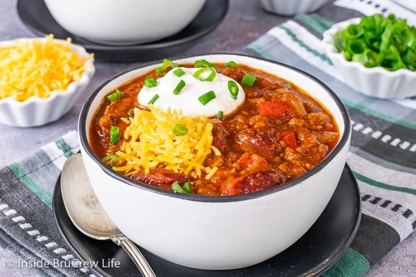 A white bowl on a black plate filled with Dr. Pepper Chili and topped with sour cream, green onions, and shredded cheddar cheese