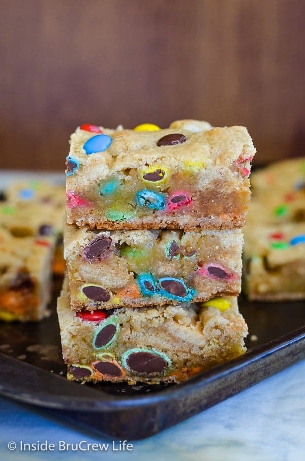A dark tray with squares of M&M Blonde Brownies on it and a stack of three in front