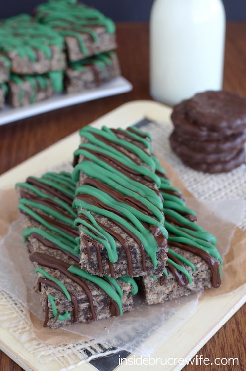 A tray with three thin mint granola bars stacked on it and a pile of thin mint cookies behind it