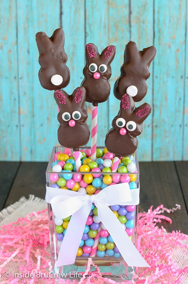 A jar of bright colored candies with a few chocolate covered marshmallow bunnies on straws in it.