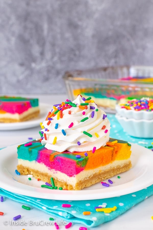 A white plate with a rainbow cheesecake bar on it topped with Cool Whip and colored sprinkles