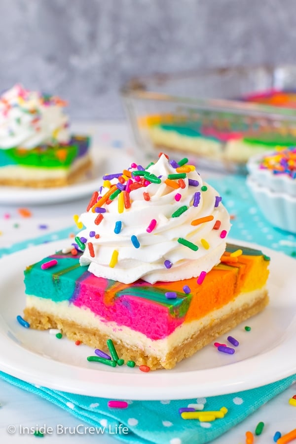 Two white plates with rainbow vanilla cheesecake bars on them with a swirl of Cool Whip and sprinkles on top