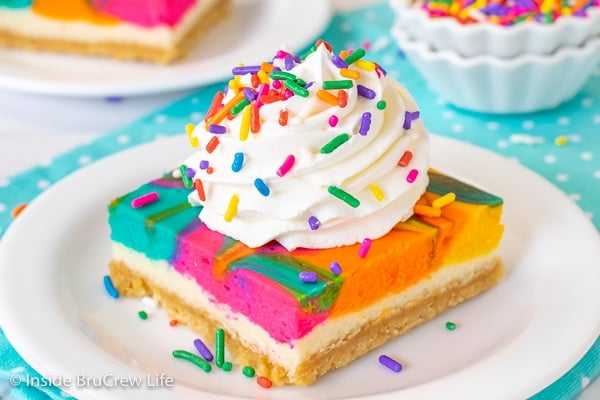 A white plate with a square of rainbow cheesecake bars topped with Cool Whip and sprinkles.