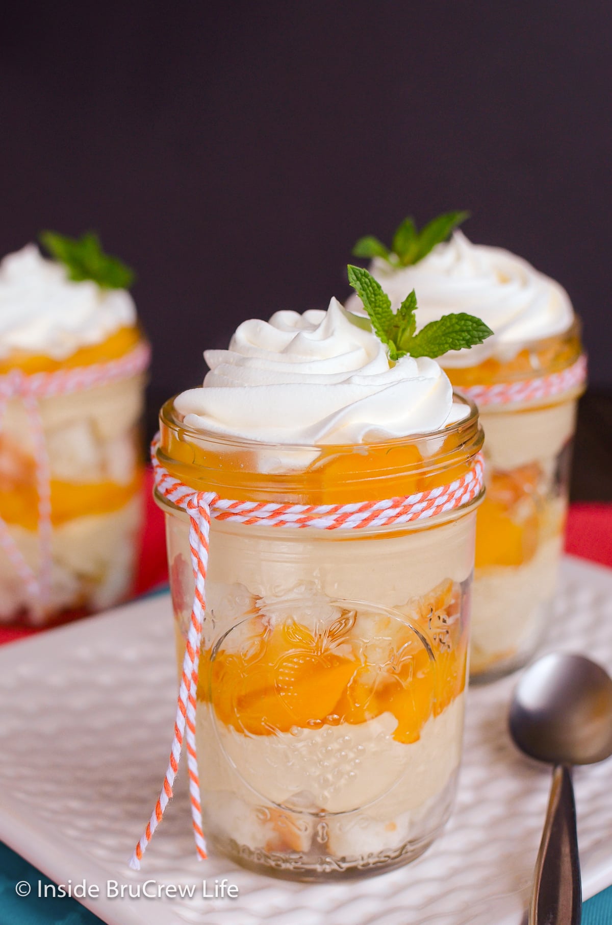 A clear jar filled with layers of cake, cheesecake, peaches, and Cool Whip.
