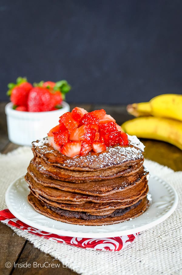 A white plate with a stack of chocolate pancakes topped with strawberries and powdered sugar on it.