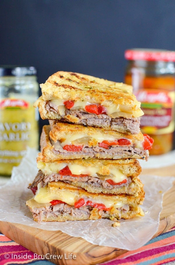 Four squares of a Cheesy Beef Panini stacked on top of each other on a cutting board with jars of ingredients behind it