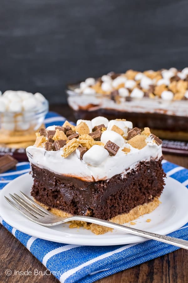 A white plate with a piece of chocolate s'mores pudding cake on it with graham crackers, chocolate bars, and marshmallows on top