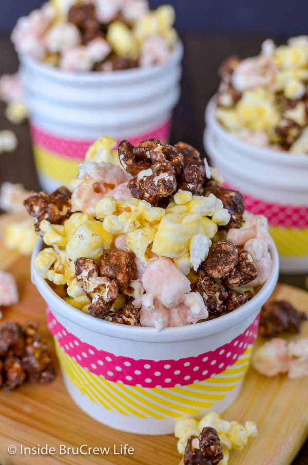 A white paper cup with pink and yellow stripes filled with banana split popcorn with more bowls behind it