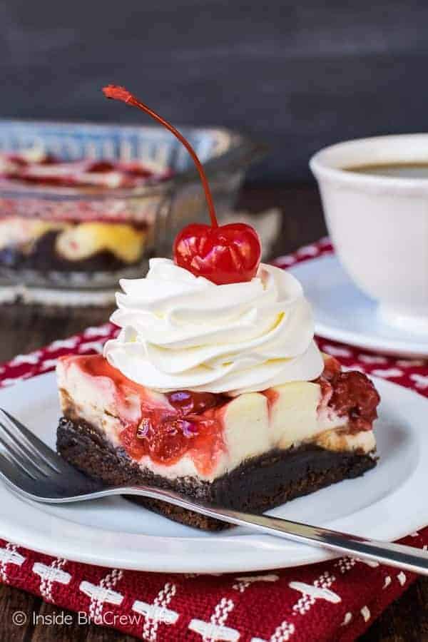 A white plate with a cherry cheesecake brownie on it topped with cool whip and a cherry