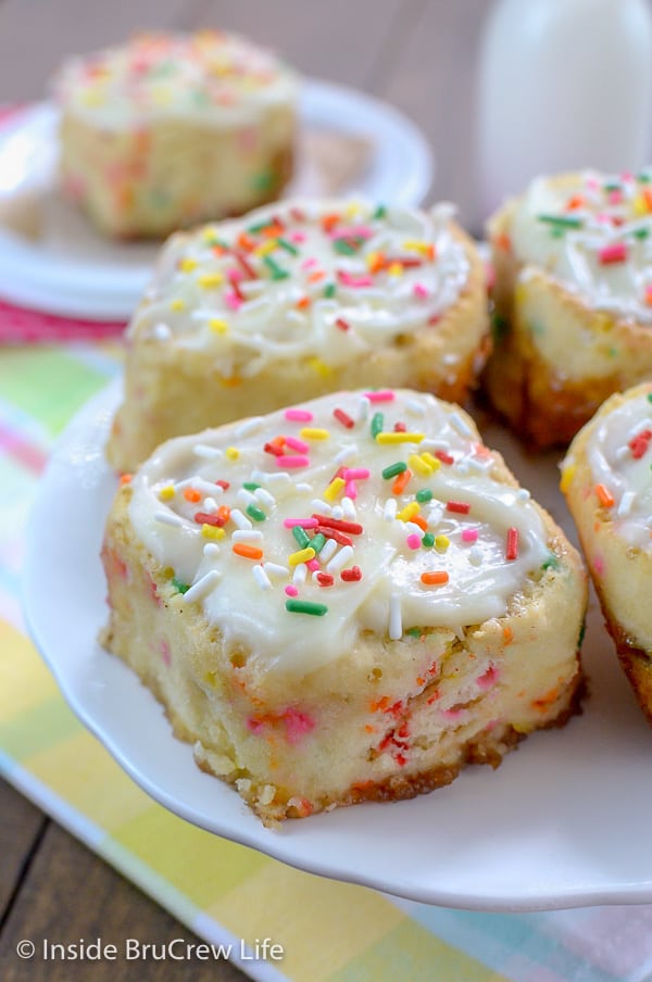 A white cake plate with frosted funfetti cinnamon rolls with sprinkles on it