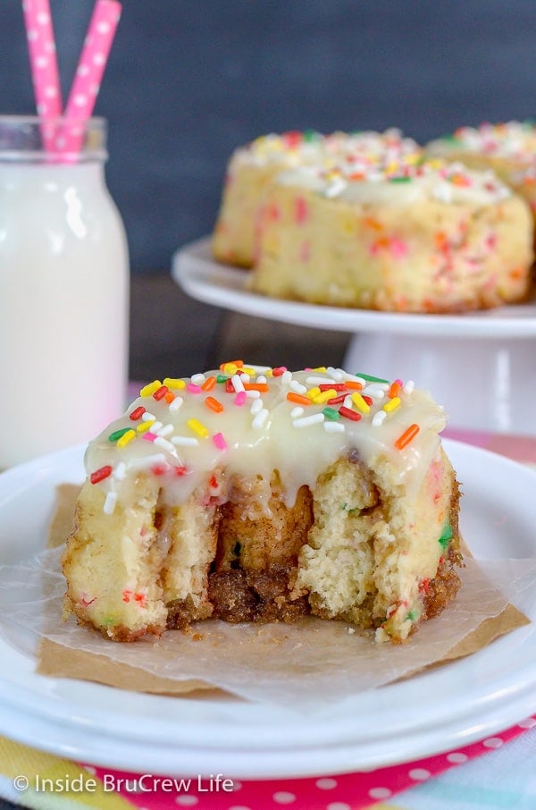 A white plate with a funfetti cinnamon roll with frosting and sprinkles on it and a bite taken out of the sweet roll