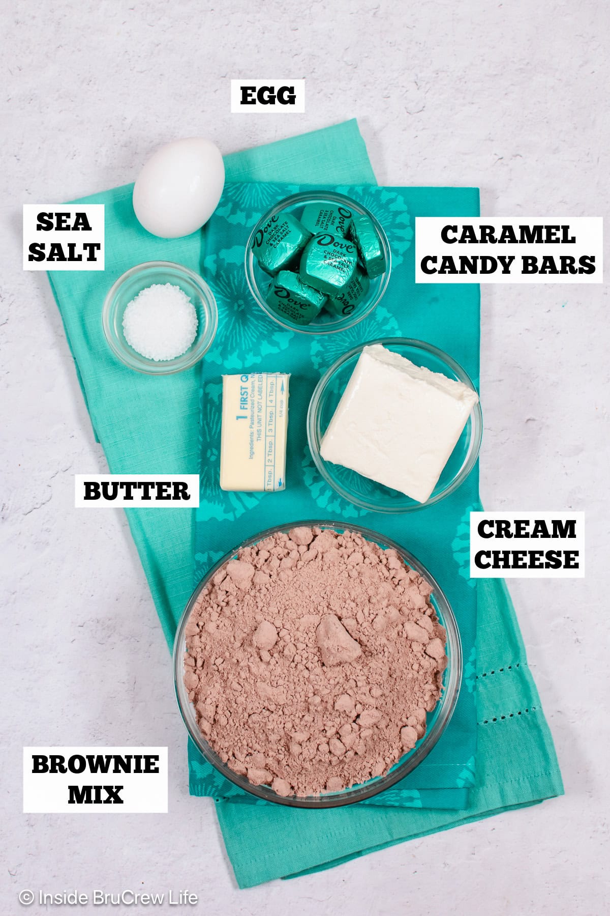 Bowls of ingredients needed to make brownie cookies with caramel bars on a white board.