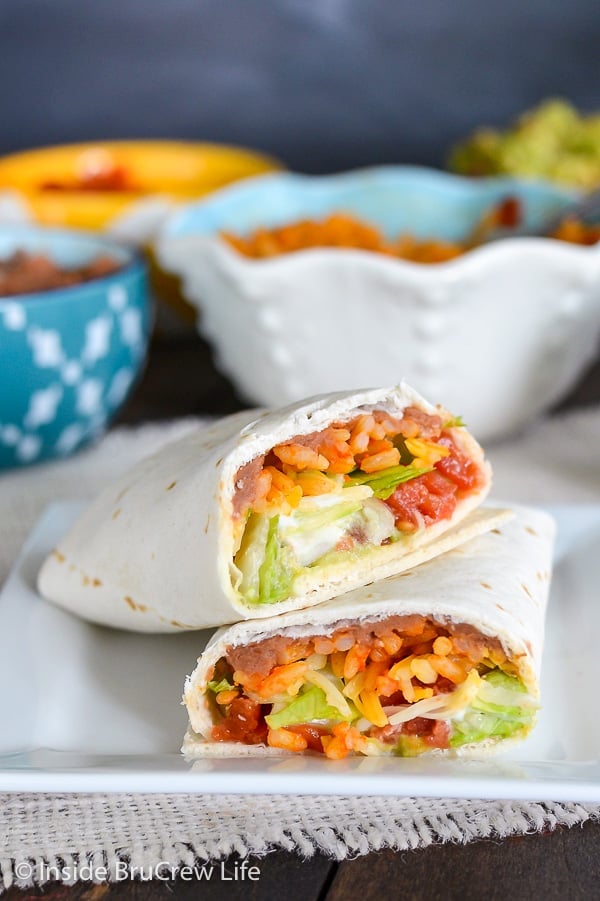 A seven layer burrito cut open on a white plate with bowls of toppings behind