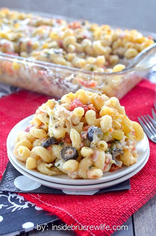 A white plate filled with cheesy veggie pasta with a pan of pasta behind it