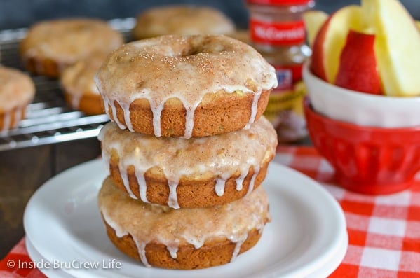 A white plate on a red and white checkered towel with three glazed cinnamon apple donuts stacked on it