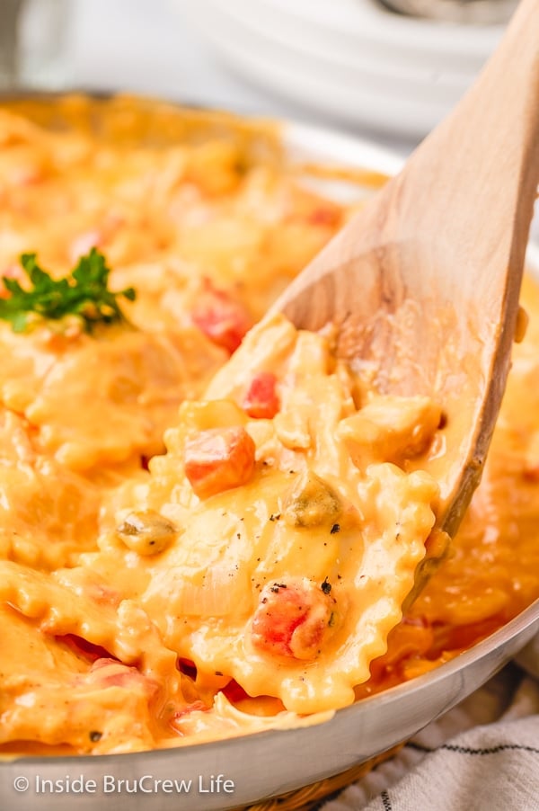 A close up picture of a wooden spoon with chicken ravioli coated with cheese sauce and tomatoes on it.
