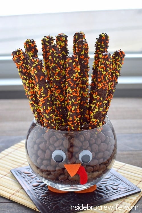 A clear jar decorated to look like a turkey and filled with chocolate covered pretzels and candies.