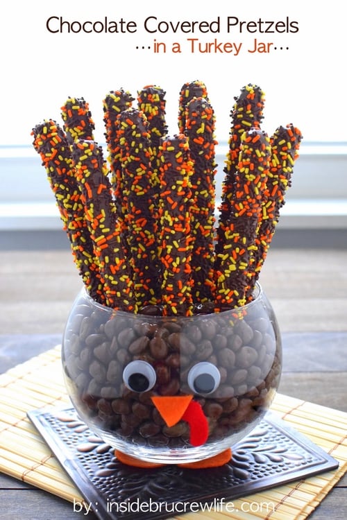 A clear jar decorated to look like a turkey filled with chocolate covered pretzels and candies.