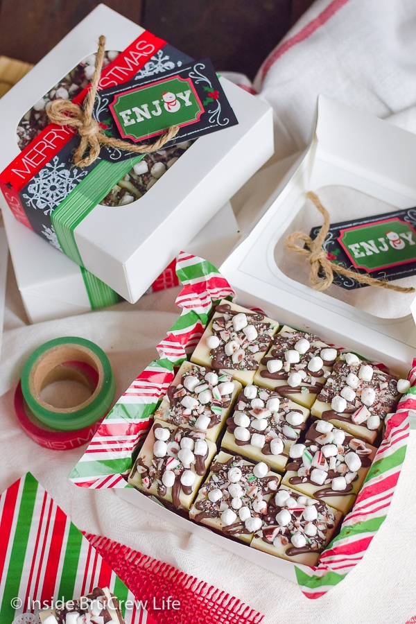 A white treat box packaged with red and green paper and pieces of candy cane hot chocolate fudge.