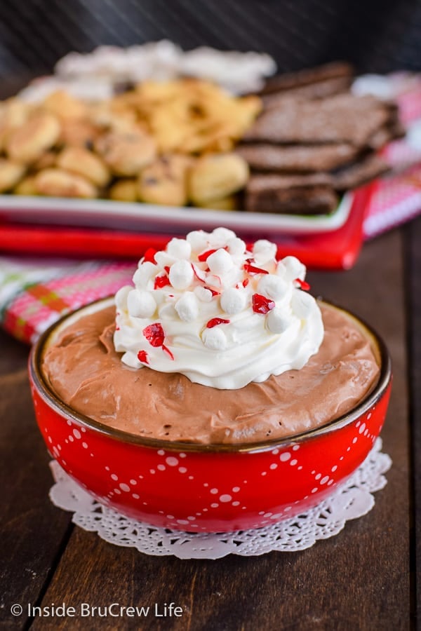 Hot chocolate cheesecake dip in a red bowl with a plate of crackers and cookies behind it.