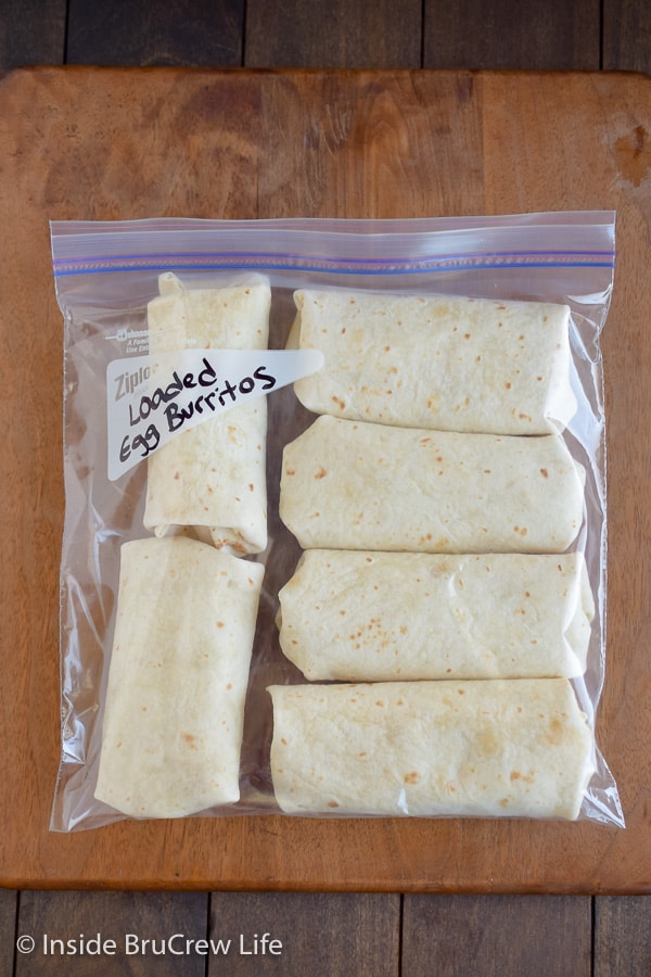 A freezer bag filled with egg burritos on a wooden cutting board.