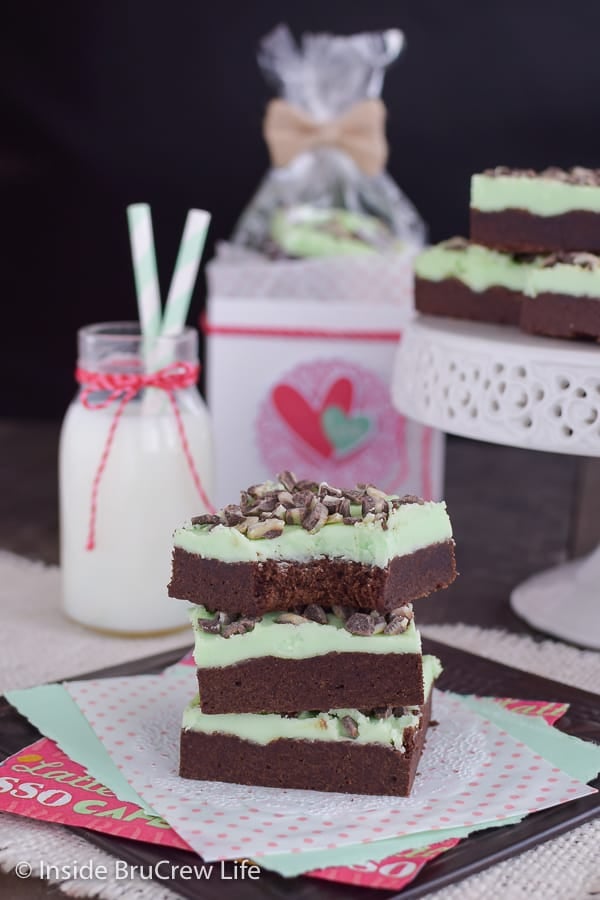 A white doily on a dark background with a stack of chocolate mint sugar cookie bars on it and milk and a tray of cookies behind it