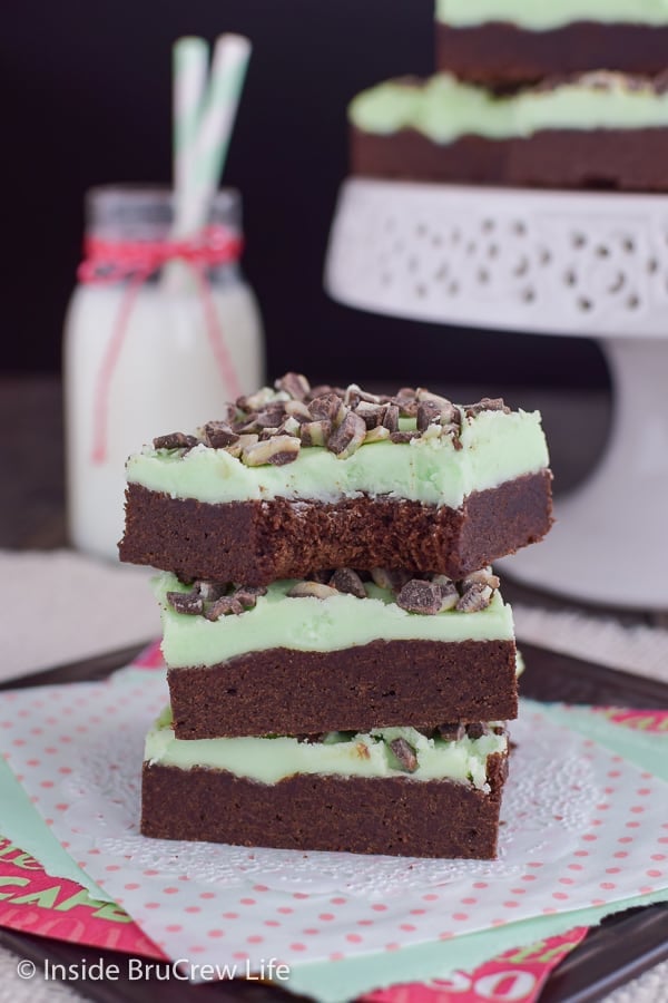 Three chocolate mint sugar cookie bars stacked together on a white doily with a tray of cookie behind it