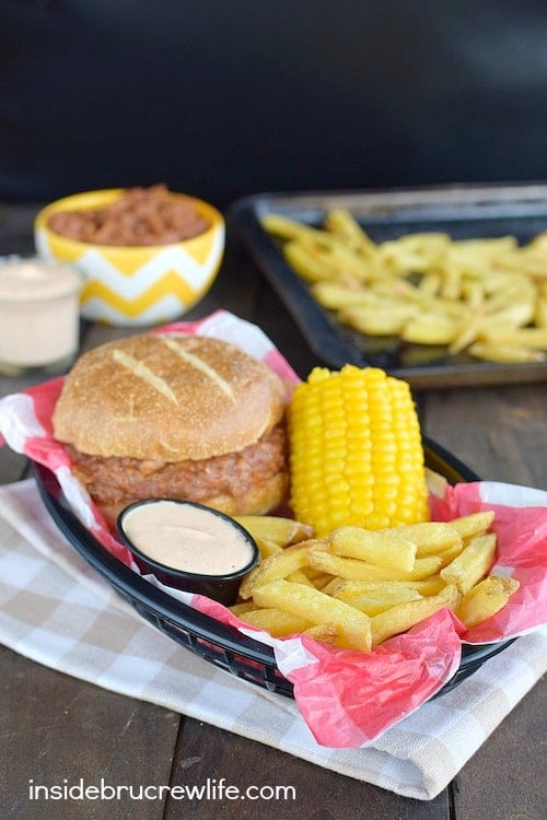 A basket with a bbq pork sandwich, fries, corn on the cob and dipping sauce.