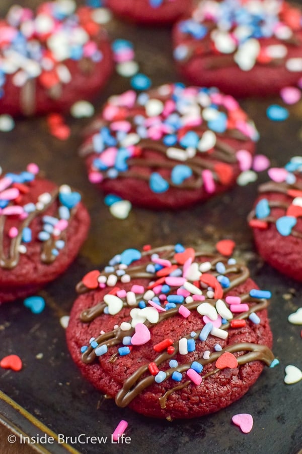 A dark cookie sheet with red velvet cookies topped with colorful sprinkles on it