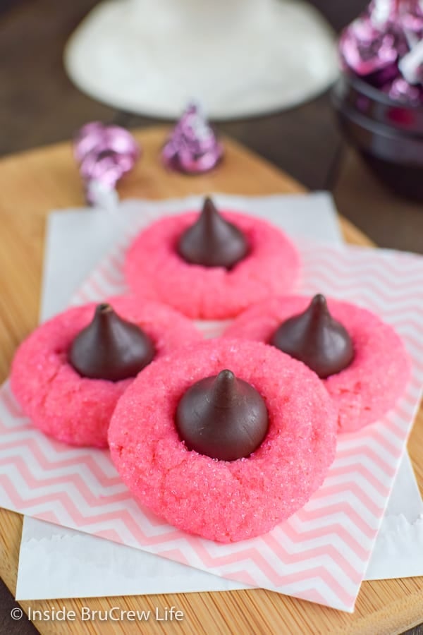 Pink cookies with chocolate kisses stacked on a wooden cutting board.