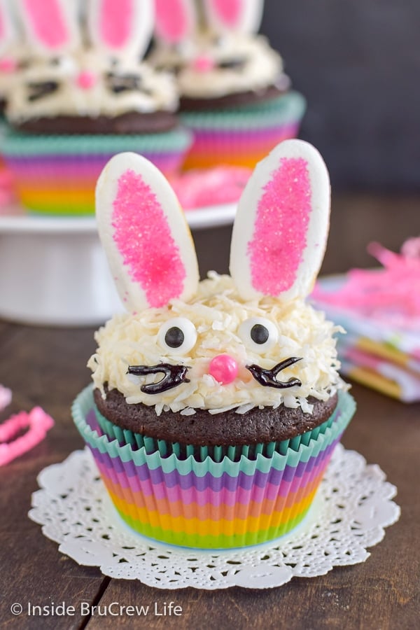 A chocolate bunny cupcake in a rainbow colored liner topped with vanilla frosting, coconut, marshmallow ears, and a candy face