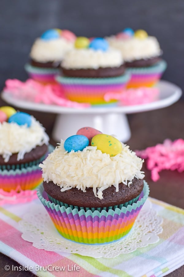 Two chocolate cupcakes topped with vanilla frosting, coconut, and candy eggs with more cupcakes behind them