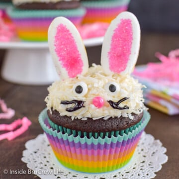 A rainbow cupcake liner with a chocolate cupcake decorated with frosting, coconut, marshmallow ears, and candy eyes