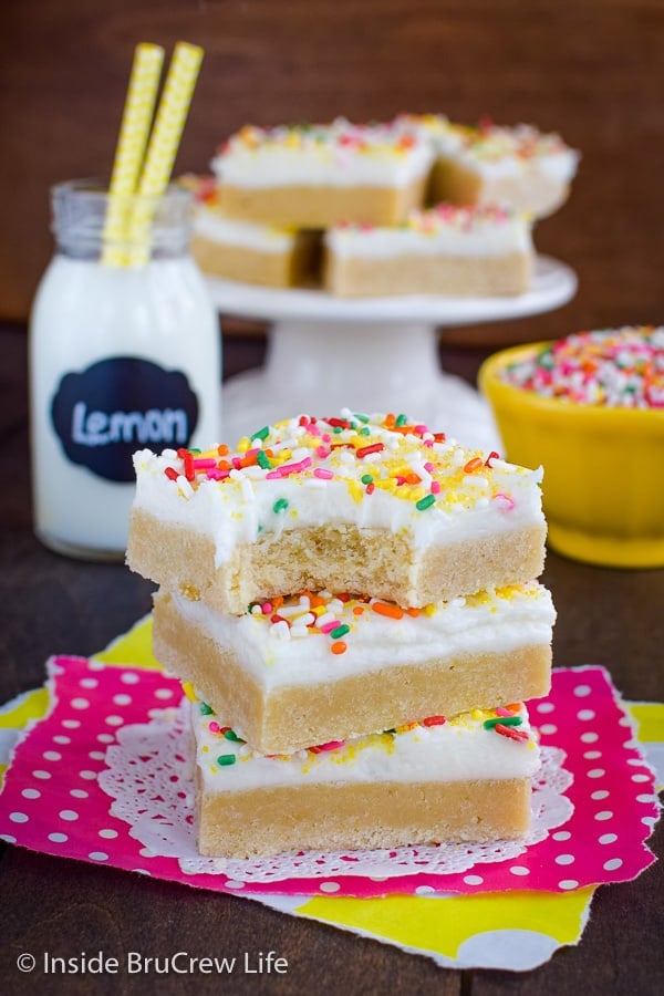 Three lemon cookie bars topped with frosting and sprinkles stacked on top of each other.