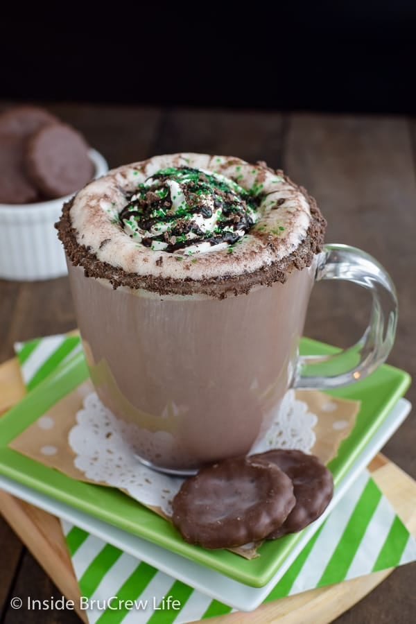 A clear coffee mug with a cookie crumble rim, chocolate syrup, cool whip, and green sugar.