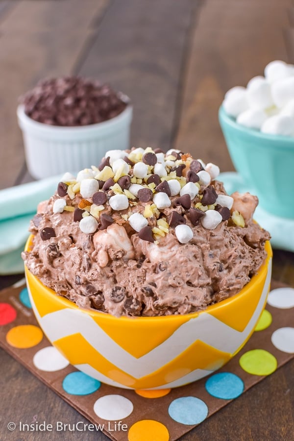 Rocky Road Fluff Salad - adding almonds, chocolate chips, and marshmallows to this easy 3 ingredient mousse makes the chocolate salad taste like rocky road ice cream. Great no bake recipe for summer picnics! A yellow and white bowl filled with chocolate fluff salad loaded with marshmallows, chocolate chips, and almond chunks