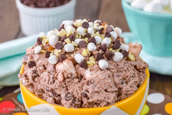 A close up picture of the top of a yellow bowl over flowing with rocky road fluff salad and extra marshmallows, chocolate chips, and almonds on top