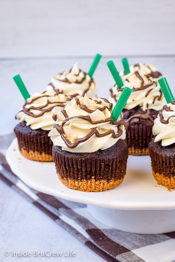 A white cake plate with frappuccino cupcakes with green straws on it.