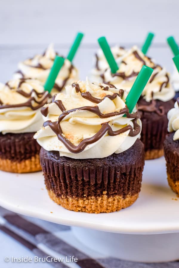 A white cake plate with chocolate cupcakes with marshmallow frosting and green straws on it.