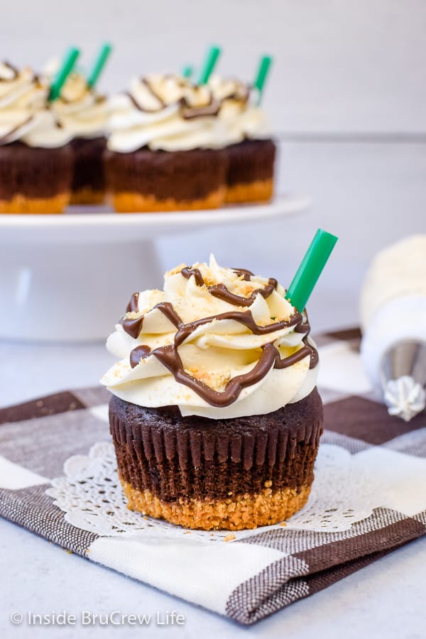 A s'mores cupcake on a white plate with a green straw in it and a cake plate behind it with more cupcakes.