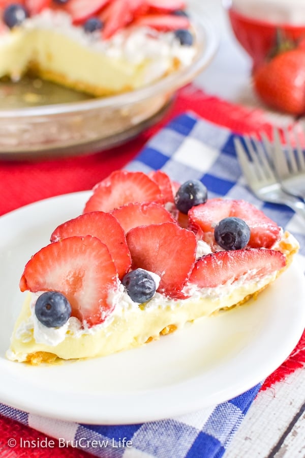 A slice of lemon cream pie topped with whipped cream, strawberries, and blueberries with the rest of the pie behind it