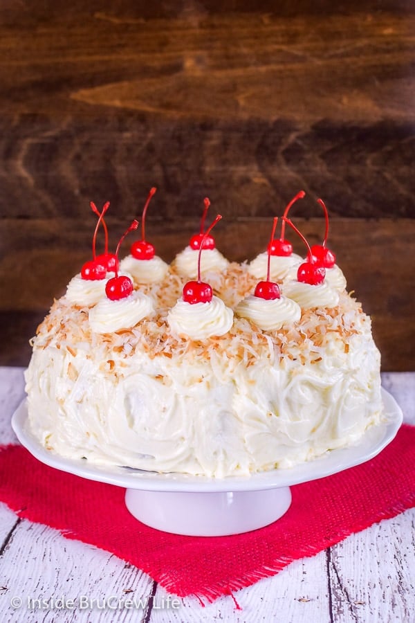 A frosted pina colada bundt cake on a white cake plate decorated with toasted coconut and maraschino cherries