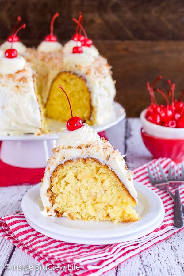 A slice of pina colada bundt cake on a white plate with a bowl of cherries and the rest of the cake behind it