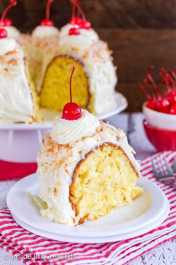 A white plate on a red and white striped towel with a slice of pina colada bundt cake on it and more cake behind it