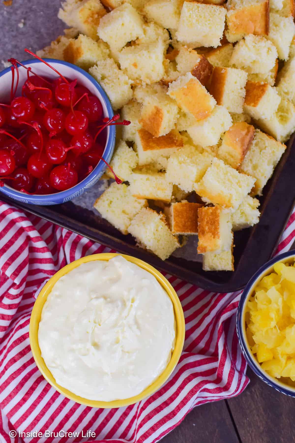 Bowls of cake, cherries, pineapple, and cheesecake filling on a board.