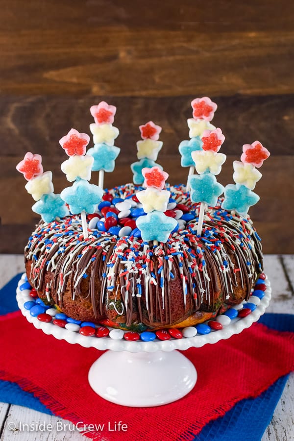 A white cake plate on a red towel with a full red white and blue bundt cake on it and marshmallow skewers all over the top