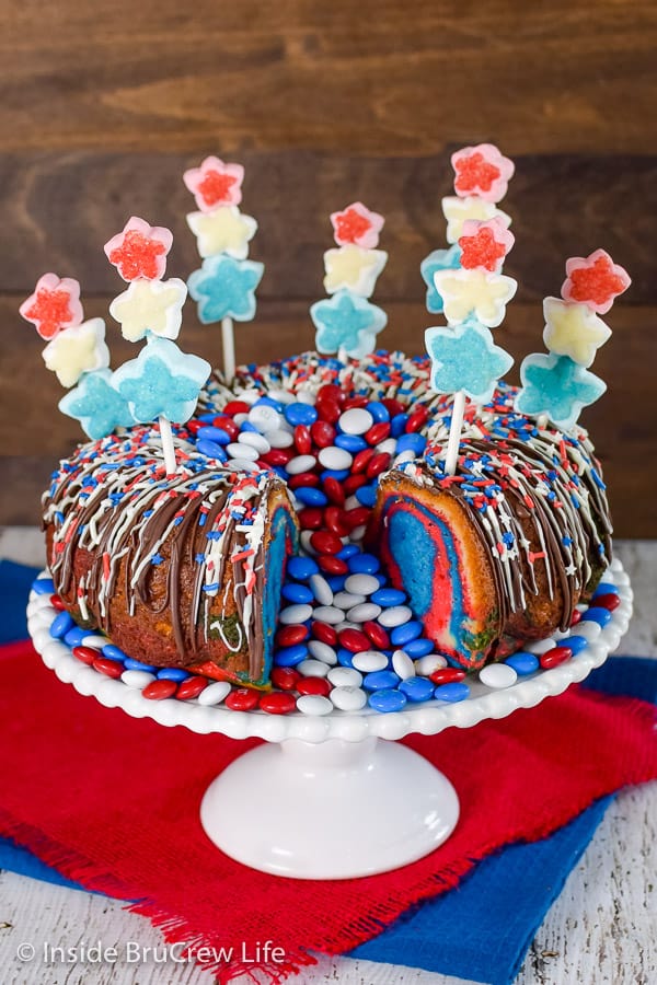 A white cake plate with the red white and blue bundt cake on it with a slice missing and red white and blue m&m's spilling out of the center