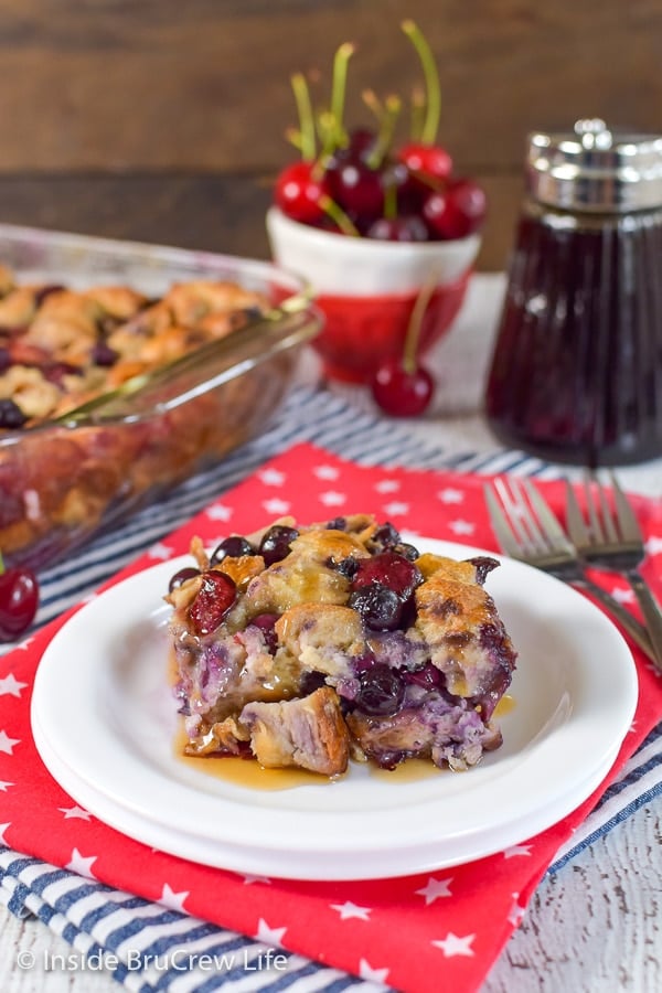 A white plate on a red towel with a square of cherry blueberry french toast bake on it and the pan behind it