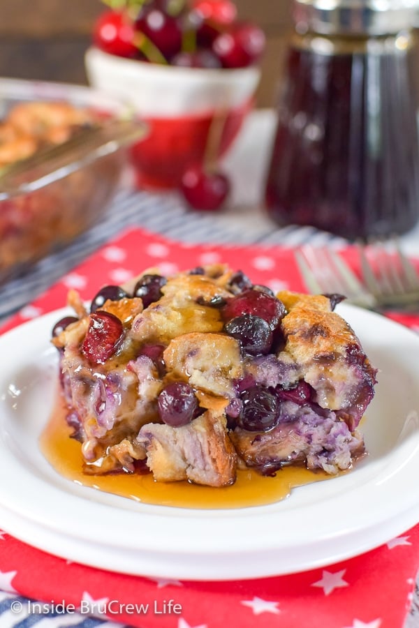 A white plate with a square of cherry blueberry french toast bake in a puddle of maple syrup